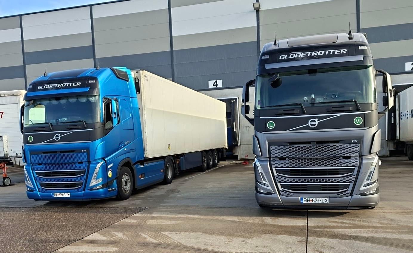 Două camioane Volvo, albastru și gri, poziționate la docuri diferite ale aceleiași hale.